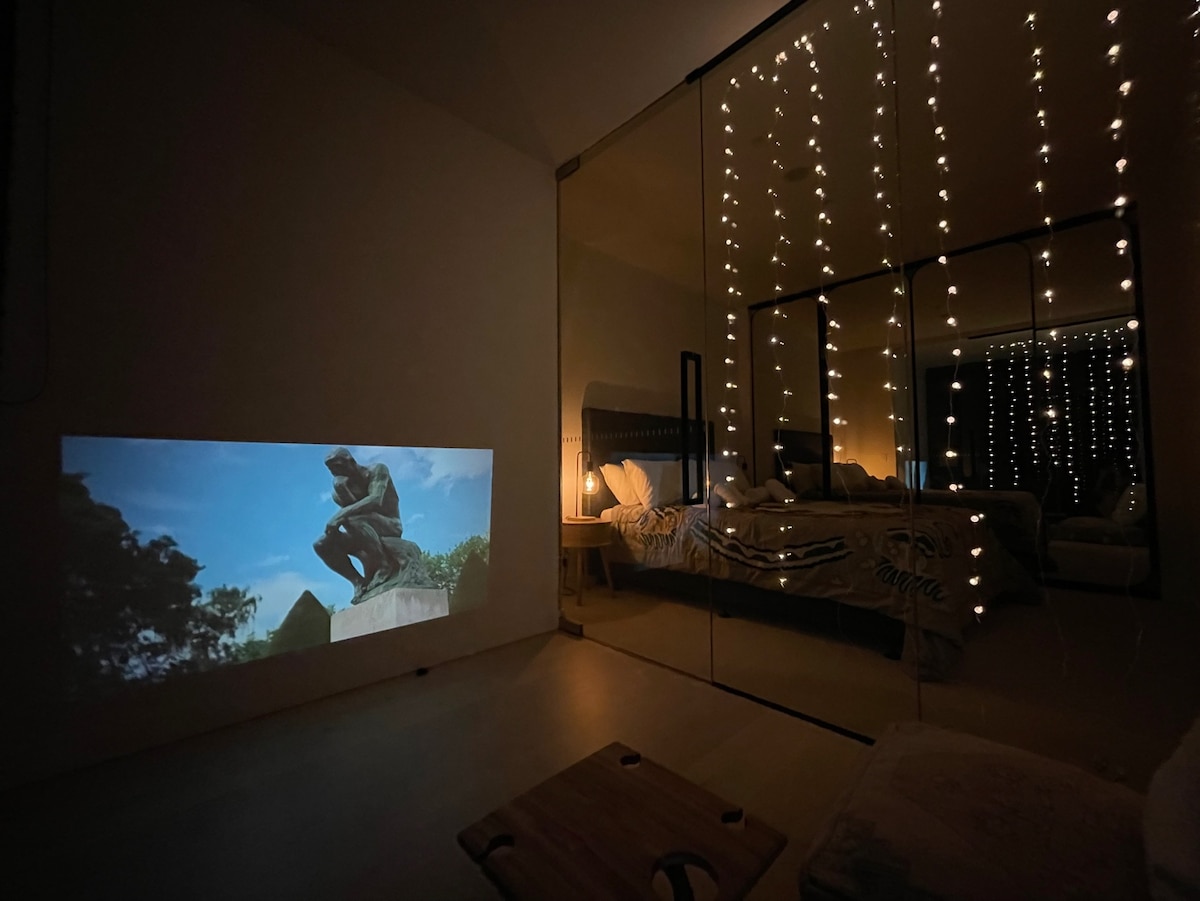 A cozy mini theatre room is showcased, featuring a projection of an image on a wall. Soft ambient lights hang above a spacious bed in a mirrored room, creating a relaxing evening atmosphere. A simple wooden tray lies on the floor, enhancing the room's comfort.