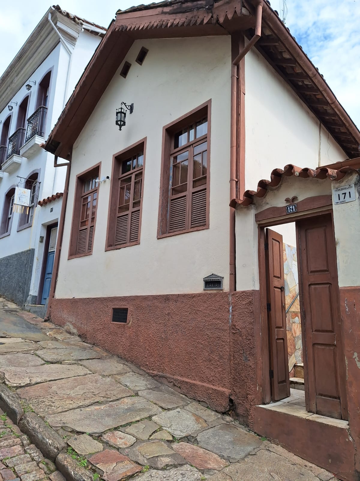 The exterior of a charming building is captured on a cobblestone street, showcasing multiple windows with wooden shutters. The entrance is framed by a rustic door, and the sloped pathway adds to the character of the location.