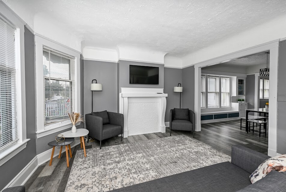 A welcoming living area features two gray armchairs flanking a circular side table. A flat-screen television is mounted above a white fireplace, with light filtering through large windows adorned with blinds. The space is enhanced by a neutral area rug against a gray wall backdrop.