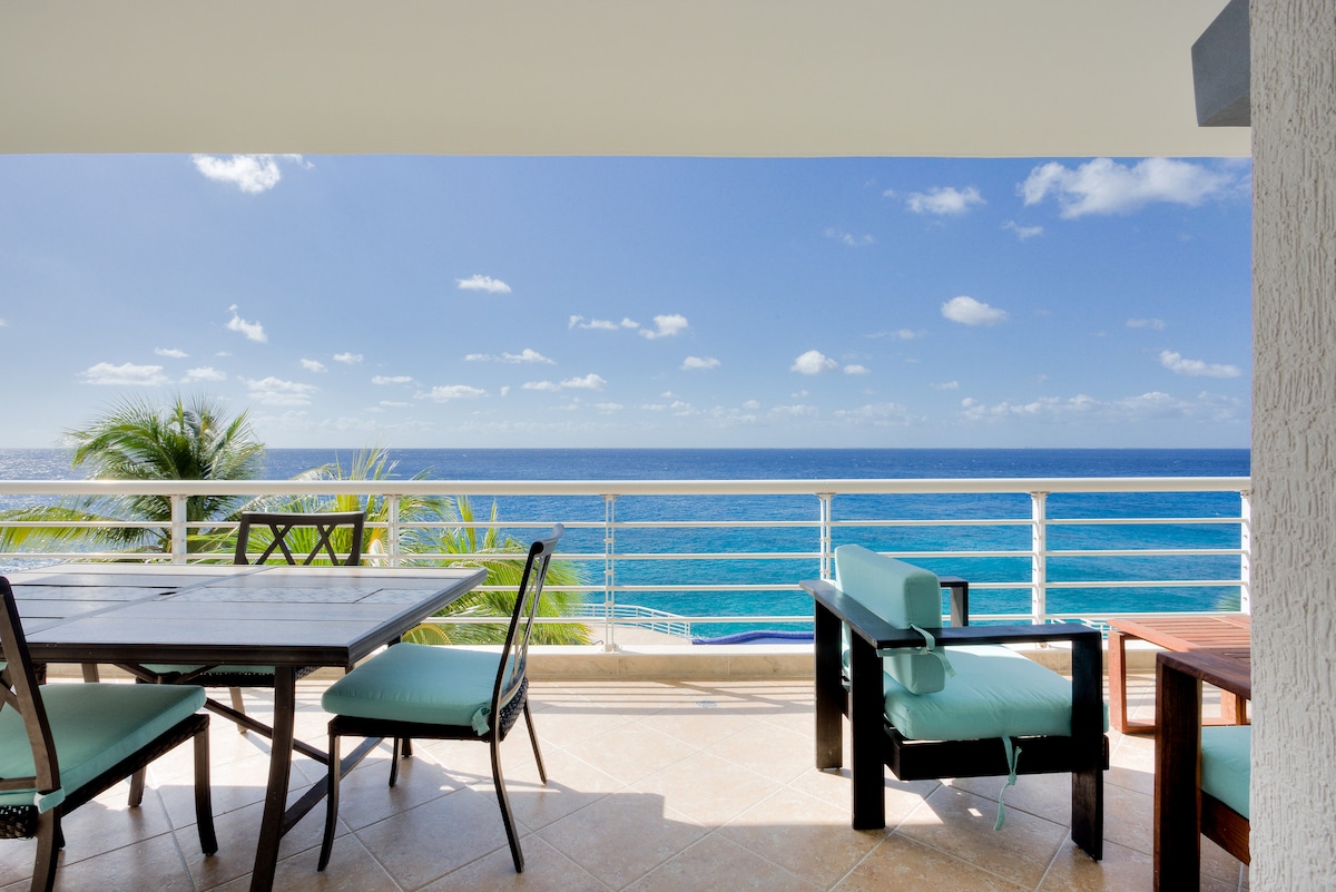 A spacious terrace is featured, overlooking the Caribbean Sea. A wooden dining table is positioned beside turquoise chairs, inviting outdoor meals with ocean views. Lush greenery is visible in the foreground, complemented by a clear blue sky and calm ocean waves in the background.