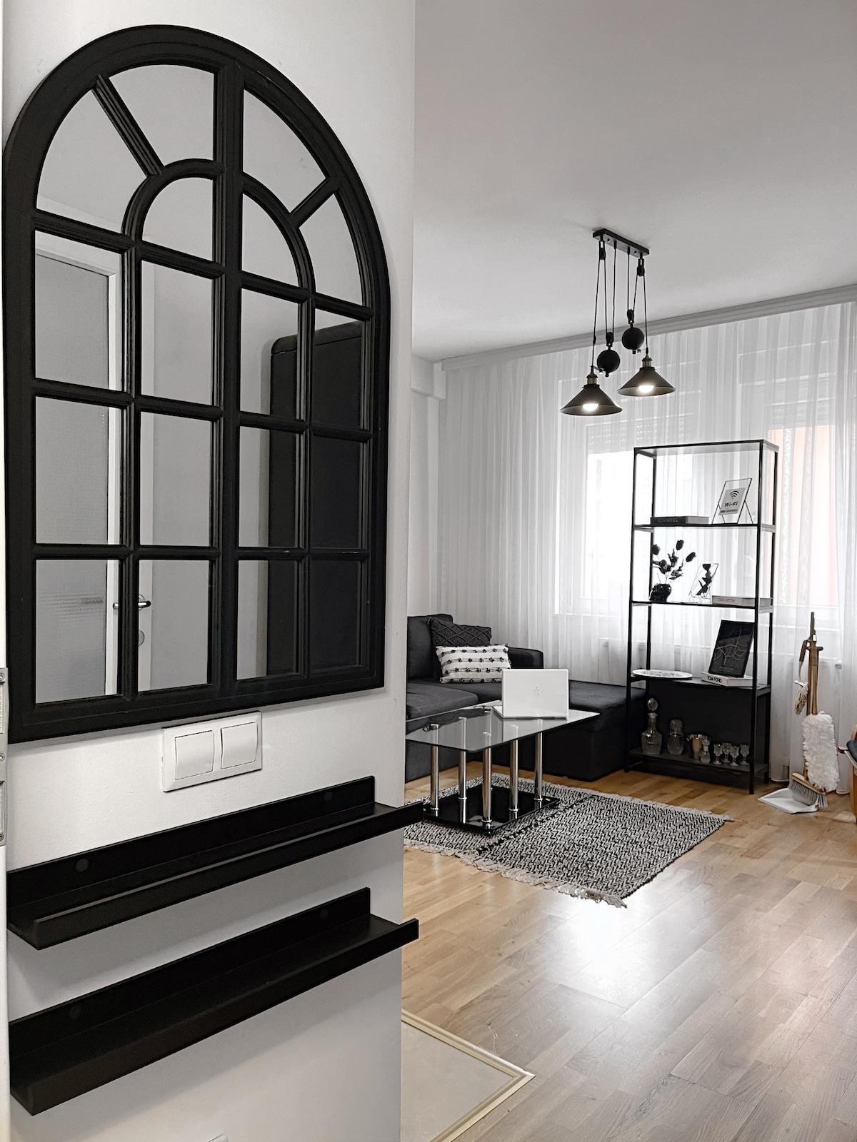 An entrance area is highlighted by a large, decorative black mirror above a sleek console with two shelves. The space opens into a living area furnished with a gray sofa, a minimalist coffee table, and a contemporary shelving unit displaying decorative items.