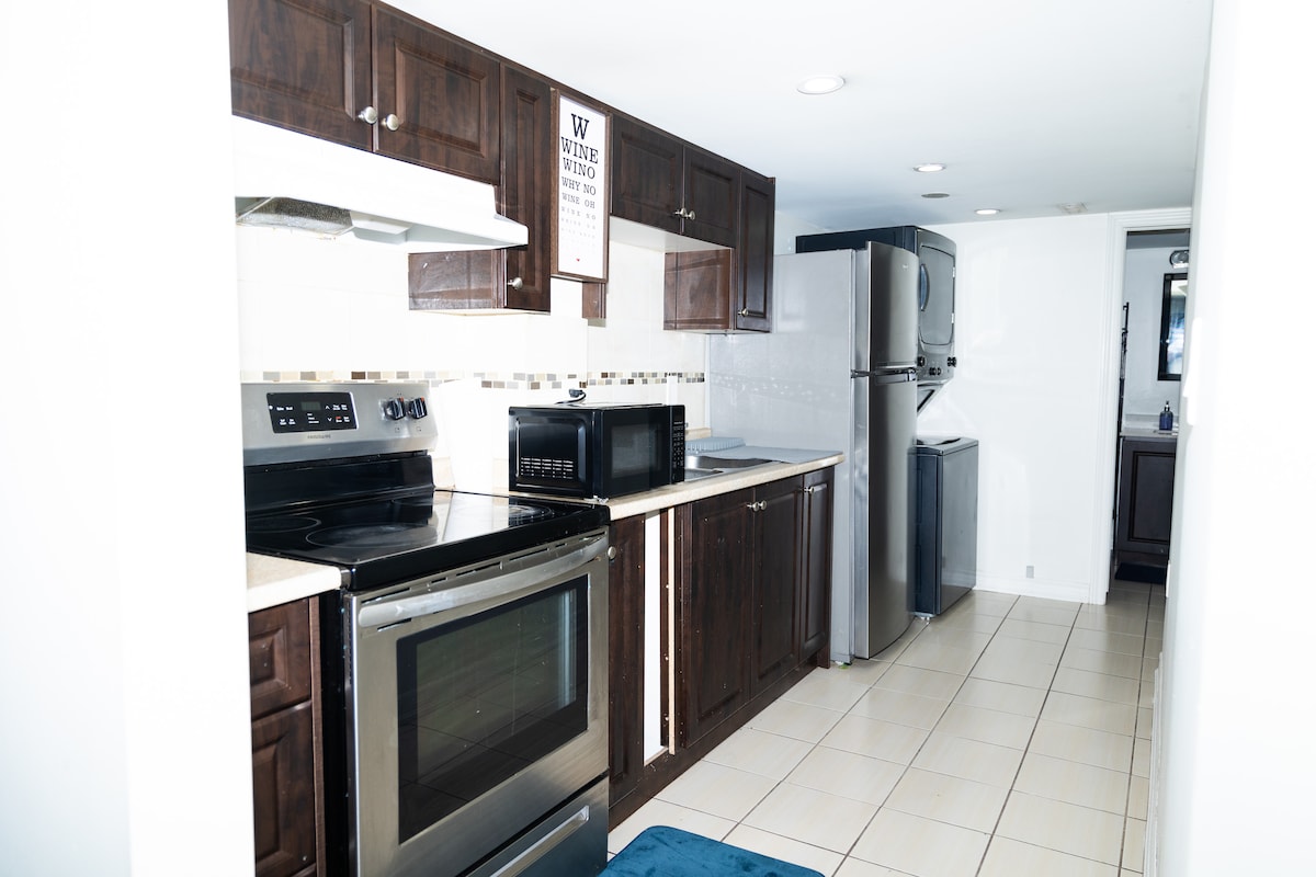 A modern kitchen featuring dark wood cabinetry and stainless steel appliances, including an oven, microwave, and refrigerator. The countertop provides ample preparation space, while white tile backsplashes enhance the overall design. A blue mat is placed on the tiled floor, adding a touch of color.