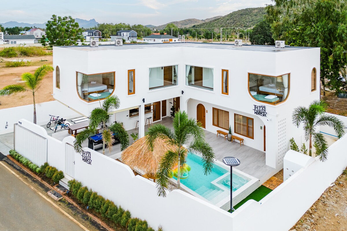 An aerial view reveals a modern two-story villa with a sleek white facade, showcasing multiple large windows. A refreshing pool is centered among palm trees and a thatched-roof gazebo in the outdoor space. An inviting terrace extends towards the lush surroundings.