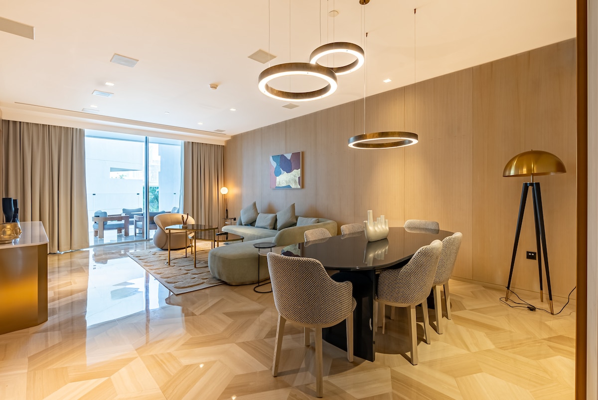 A modern dining area features a sleek table surrounded by textured chairs, complemented by a contemporary light fixture above. A spacious living area showcases a large sectional sofa and a stylish lounge chair, with floor-to-ceiling windows leading to an outdoor terrace.