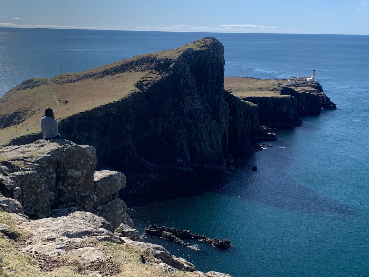 A coastal landscape is showcased, featuring rugged cliffs that descend into the calm blue waters. In the distance, a lighthouse stands prominently on the horizon, while the green hills gently recede into the background under a clear sky.