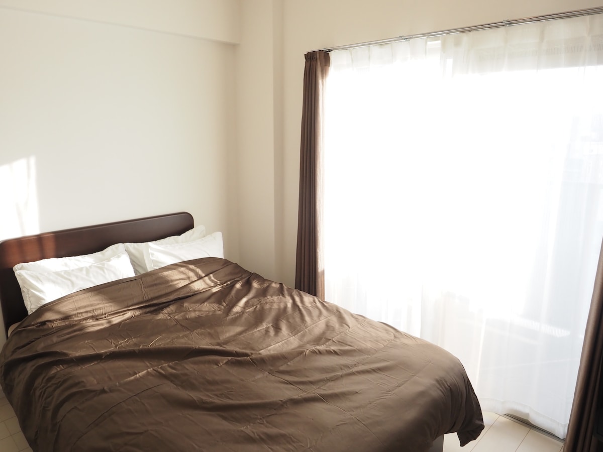A spacious bed is presented with a cozy brown coverlet, surrounded by light-colored walls. Natural light filters through sheer white curtains, creating a soft ambiance in the room. The window provides a view of the outside space while maintaining privacy.