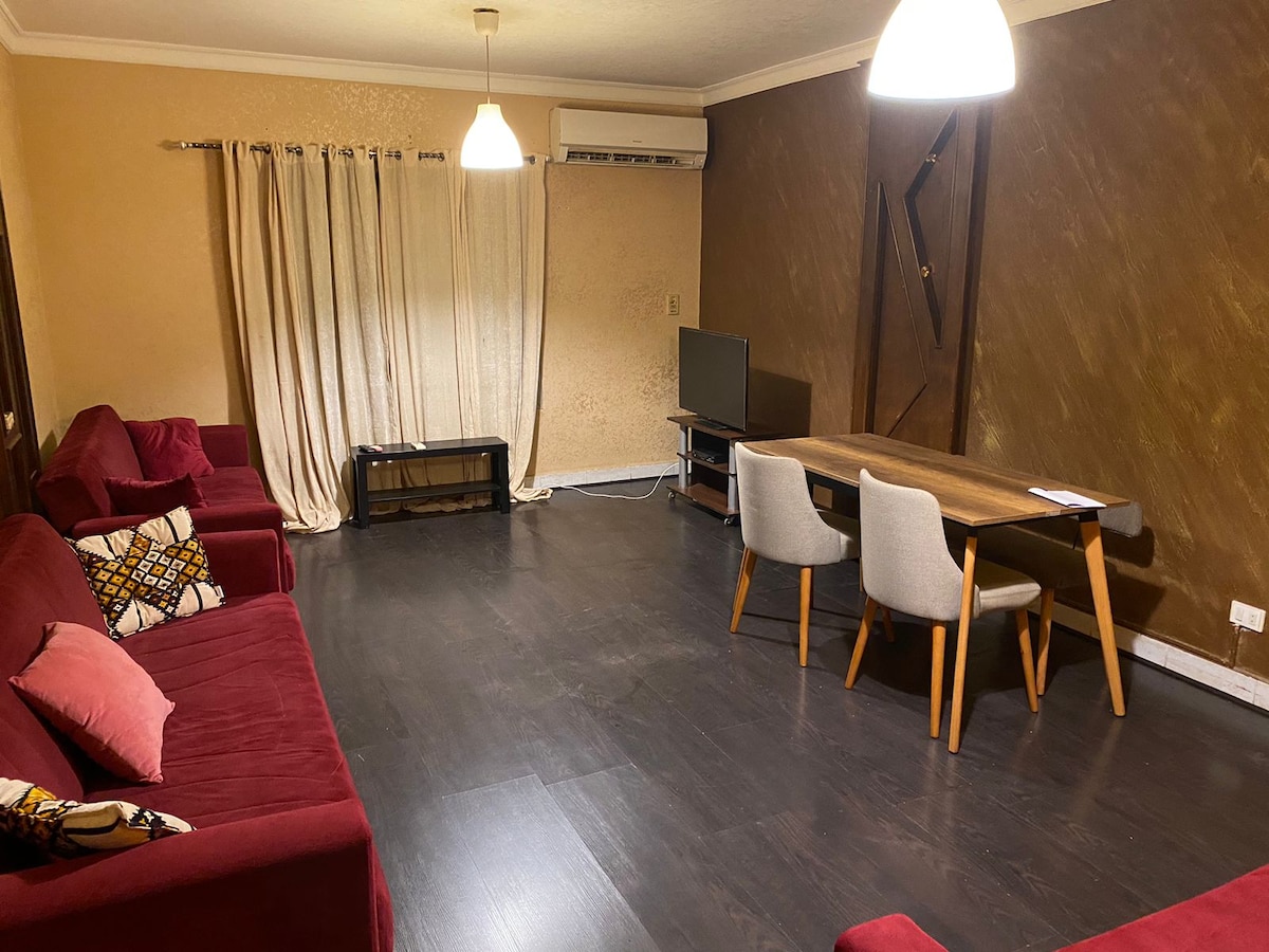 The reception area is furnished with three red sofas and a dining table surrounded by chairs. A television is positioned on a low stand, and curtains frame the large window. Soft overhead lighting complements the warm wall tones.