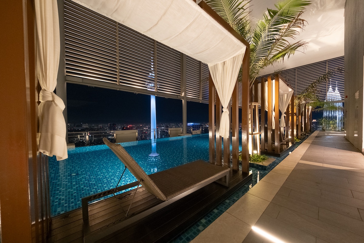 A rooftop area features a sleek pool illuminated by soft lighting, bordered by elegant palm trees. Comfortable loungers are situated beside the water, offering views of the surrounding city skyline. The space is designed for relaxation and enjoyment, with a tranquil atmosphere created by the serene night setting.