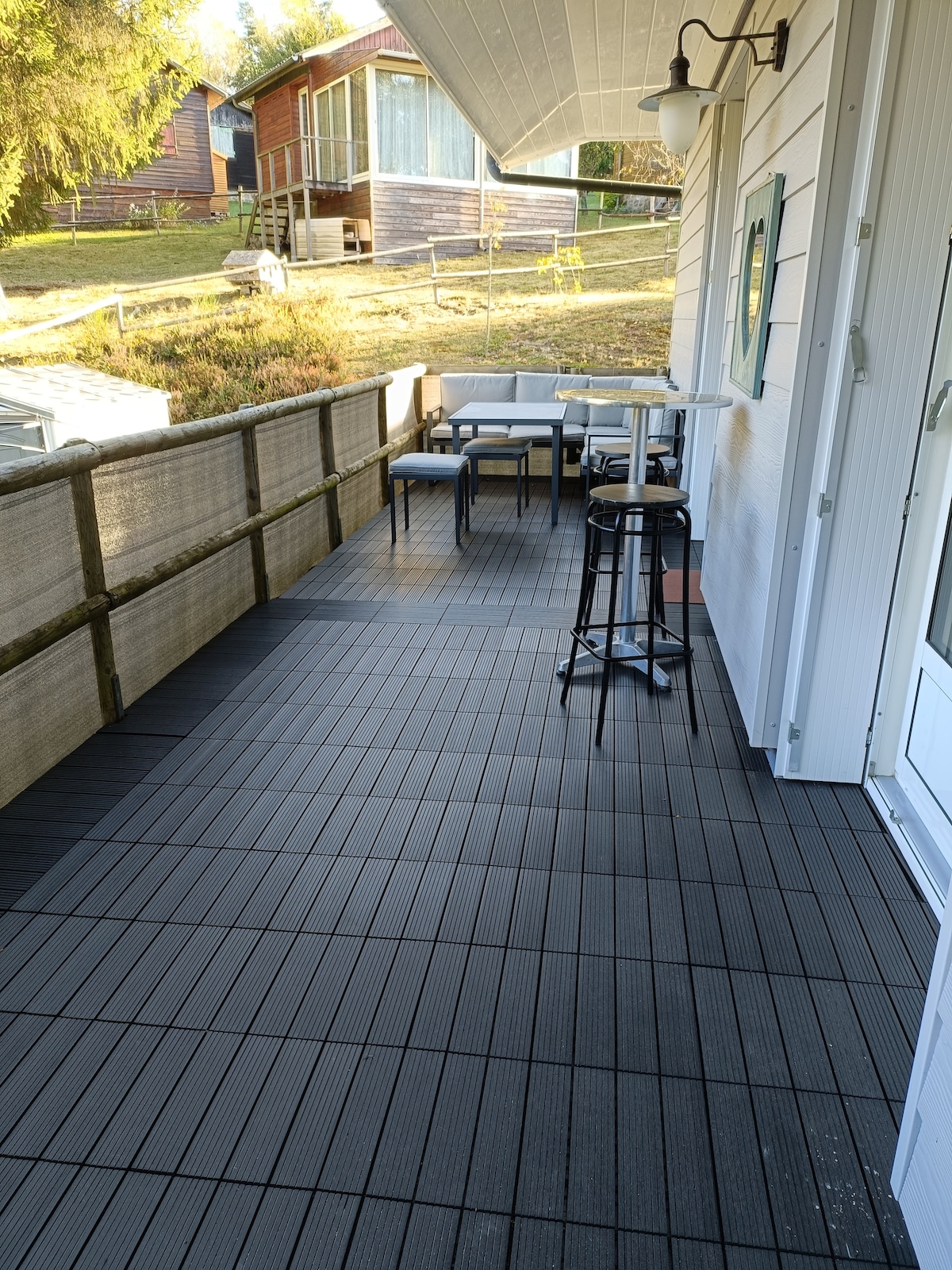 A flat terrace area features dark composite decking. A small table and two chairs are arranged for outdoor dining. A high barstool is positioned near the railing, providing a space for relaxed seating. Nearby cabins are visible in the background.