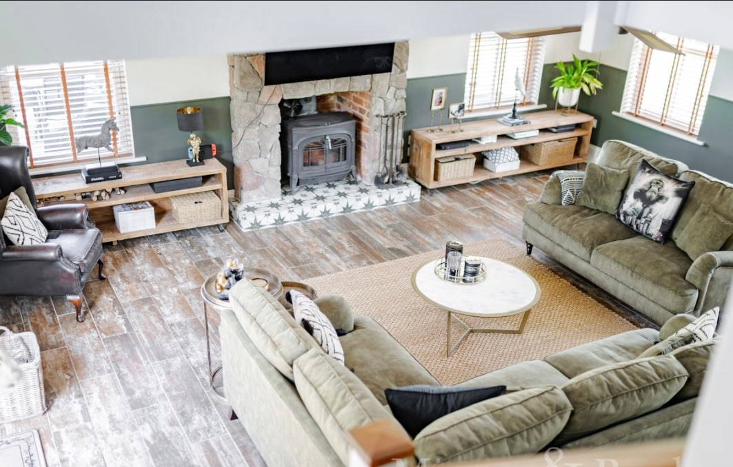 An open-plan living area showcases a cozy seating arrangement with two green sofas and a leather armchair. A round coffee table sits in the center on a jute rug, while natural light filters through the plantation shutters. A stone fireplace adds warmth, surrounded by wooden shelves.