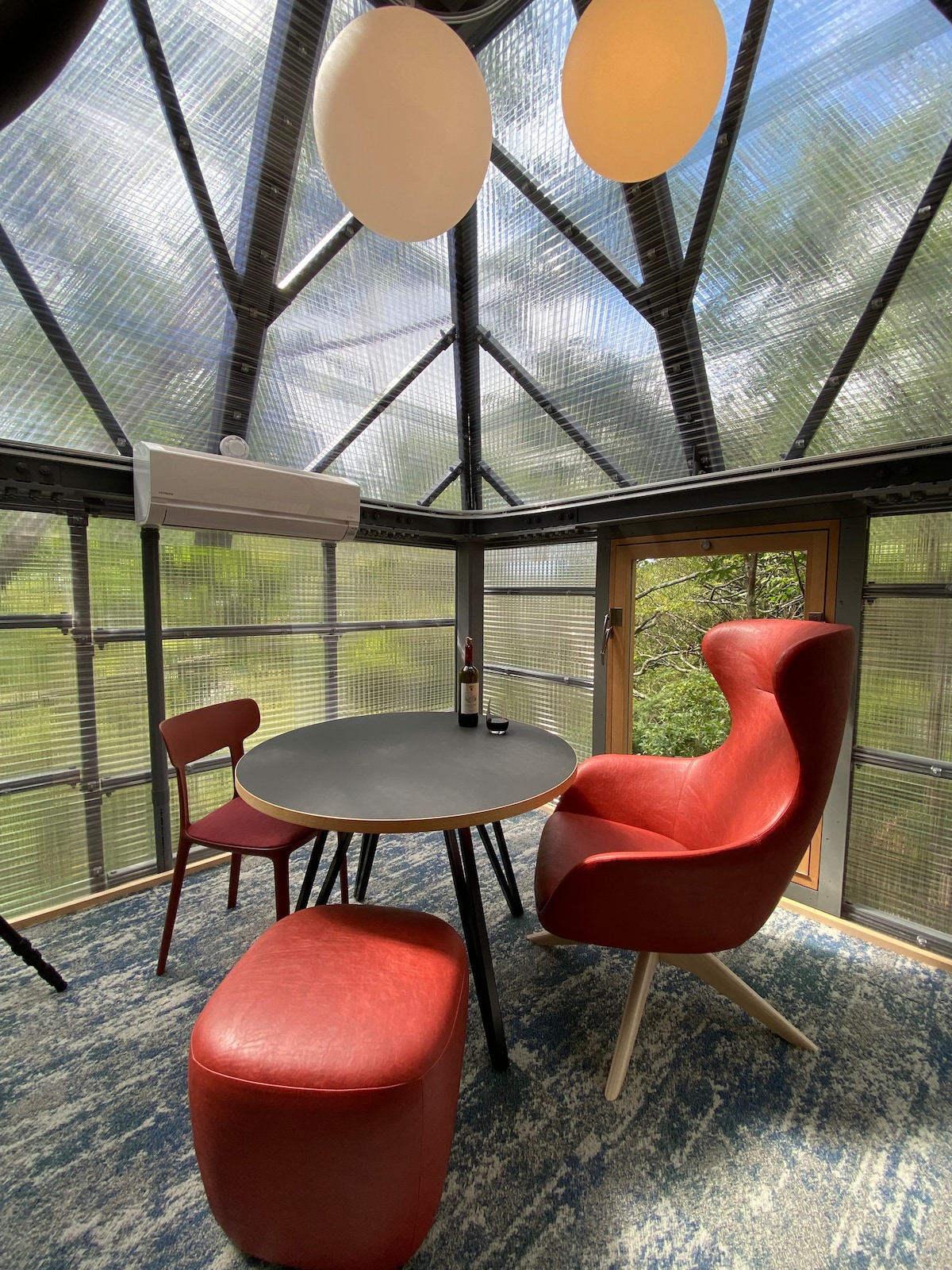 A bright space features a circular table surrounded by two red chairs and a contemporary armchair. Natural light streams in through a triangular glass roof, highlighting the green outdoors visible through the windows. A small bottle of wine rests on the table.