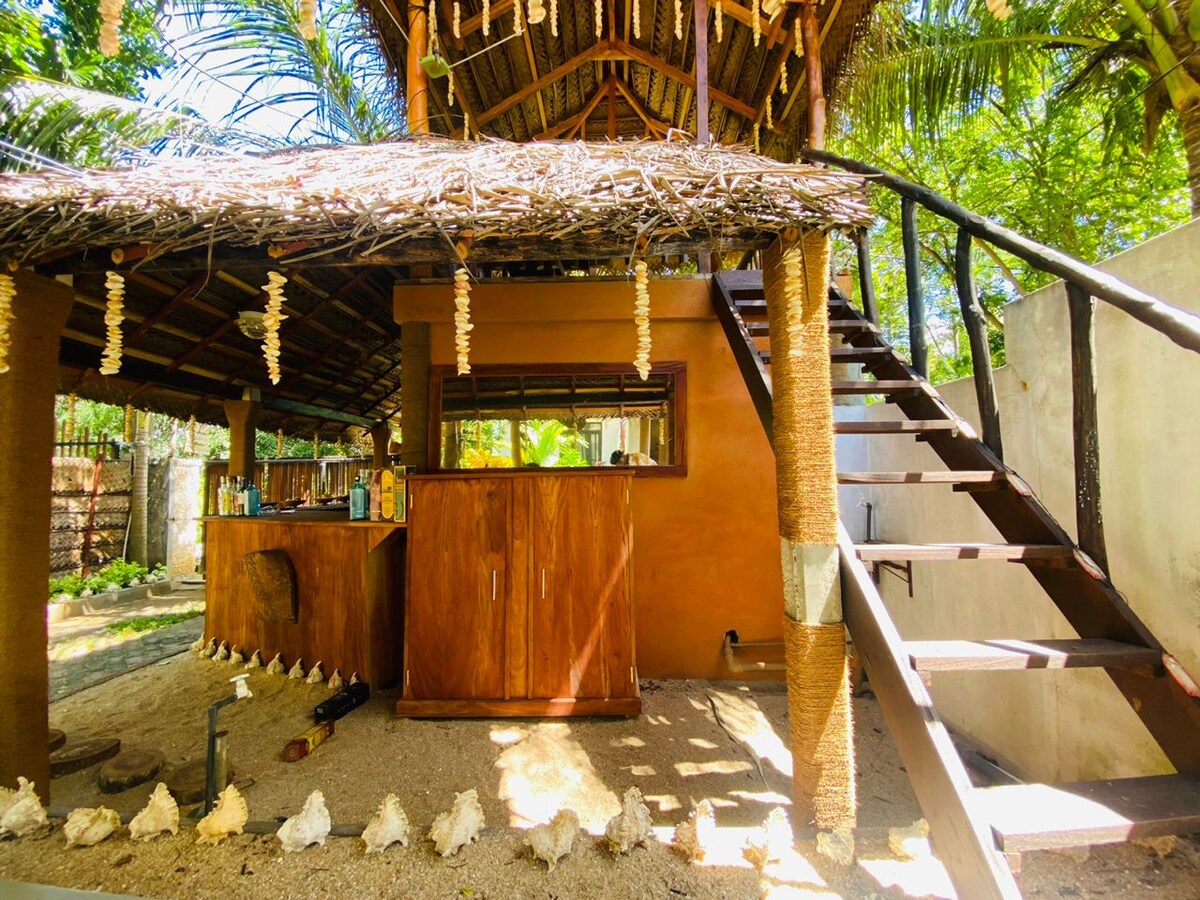 A rustic outdoor bar area is visible beneath a thatched roof, with wooden cabinets arranged neatly. Stairs lead to an upper level, surrounded by lush greenery. The ground is adorned with small shells, adding a natural element to the inviting space.