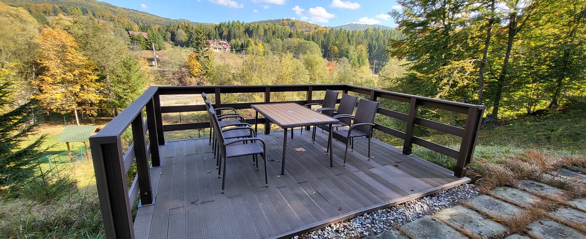 An outdoor terrace features a large wooden table surrounded by six chairs, set against a backdrop of lush green hills and autumn trees. The area is enclosed by a wooden railing, providing a serene space for relaxation.