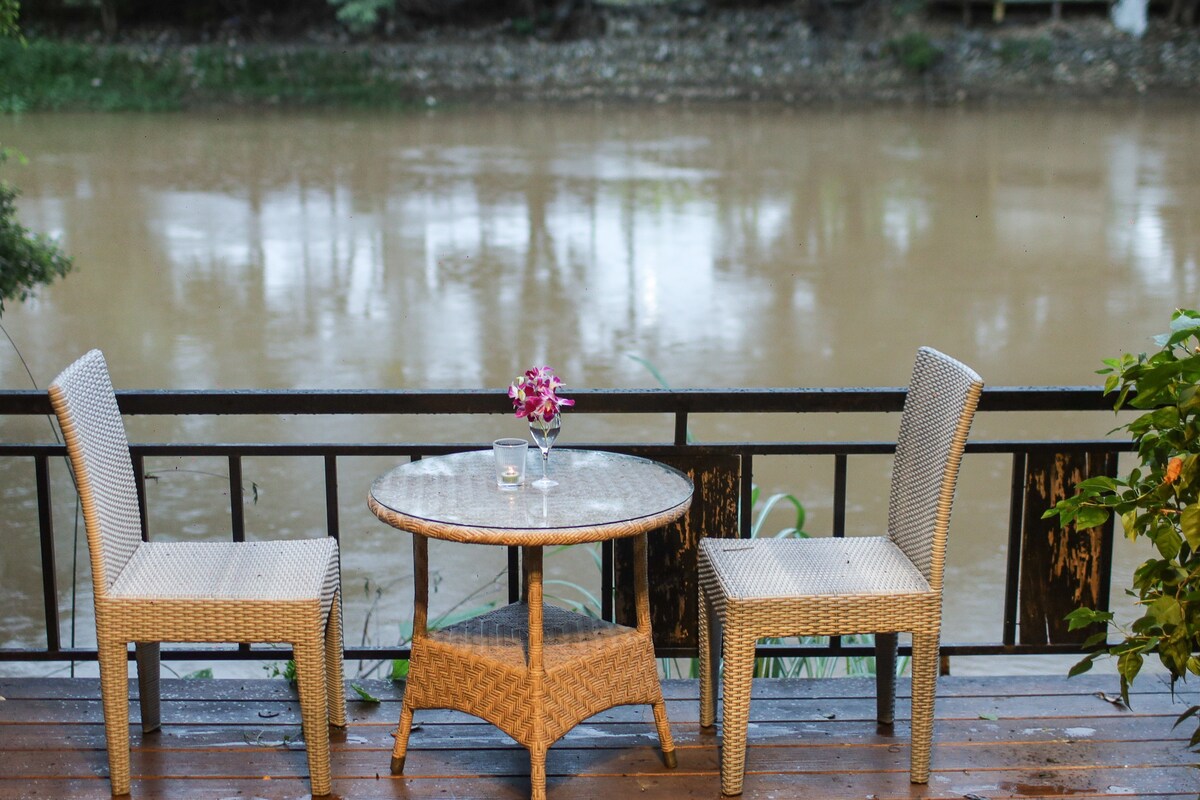 A small outdoor seating area features a round wicker table accompanied by two chairs, positioned beside the tranquil Ping River. A vase with an orchid sits on the table, enhancing the natural setting with simple elegance.
