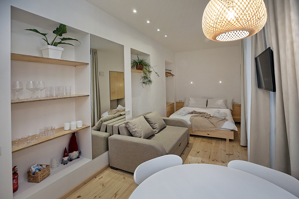 A modern living area features a light-colored sofa adjacent to a spacious bed with soft linens. Open shelving displays glassware and decorative items. A round dining table is positioned in the foreground, and plants add a touch of greenery, while soft lighting enhances the serene atmosphere.