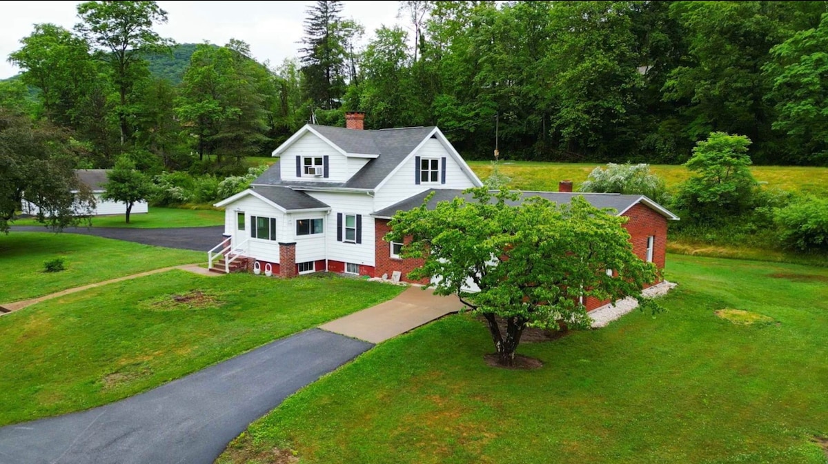 The exterior of the lodge features a two-story structure with a mix of white siding and red brick. A manicured lawn surrounds the home, enhanced by green trees. A paved driveway leads to the entrance, with steps ascending to the front porch.