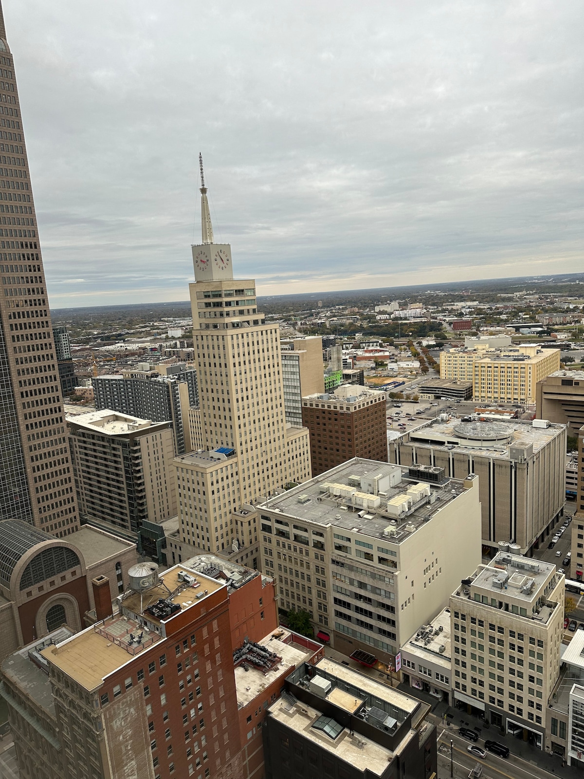 A cityscape is presented from a high vantage point, featuring a mix of architectural styles. Distinguished buildings, including a tower with a clock, are visible among a landscape of urban structures and rooftops. The scene is set against a cloudy sky, creating a muted atmosphere.