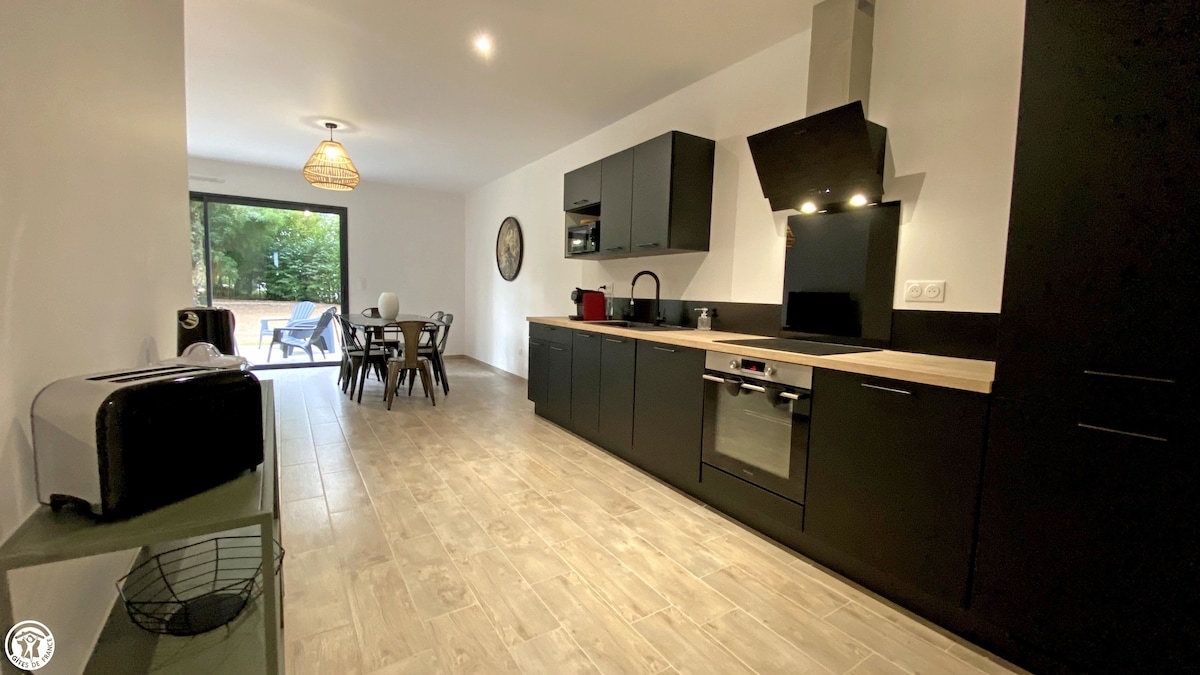 A modern kitchen area features sleek black cabinetry and a light-colored countertop. An open layout connects to a dining space with a circular table and chairs. Large glass doors lead to a patio, allowing natural light to brighten the room.