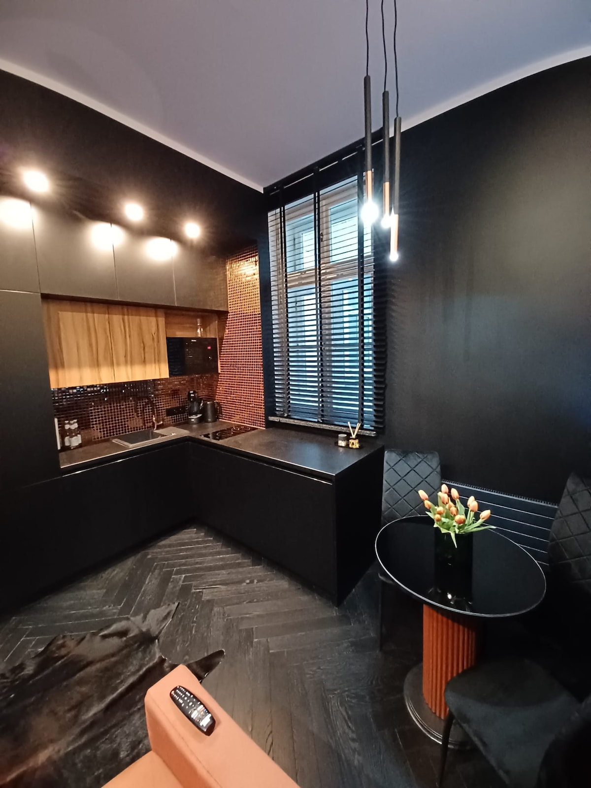 A modern kitchenette is highlighted with dark cabinetry and warm wooden accents. A small, round table with a floral centerpiece sits beside plush seating, while a window covered with horizontal blinds allows soft light to filter in, creating a cozy and intimate setting.