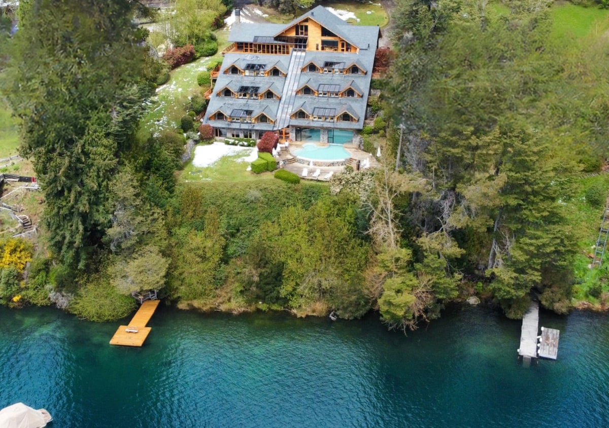 The hotel is nestled on a verdant hillside overlooking a calm lake. The structure features multiple levels with large windows, surrounded by lush greenery. A private dock extends into the water, complementing the serene natural setting.