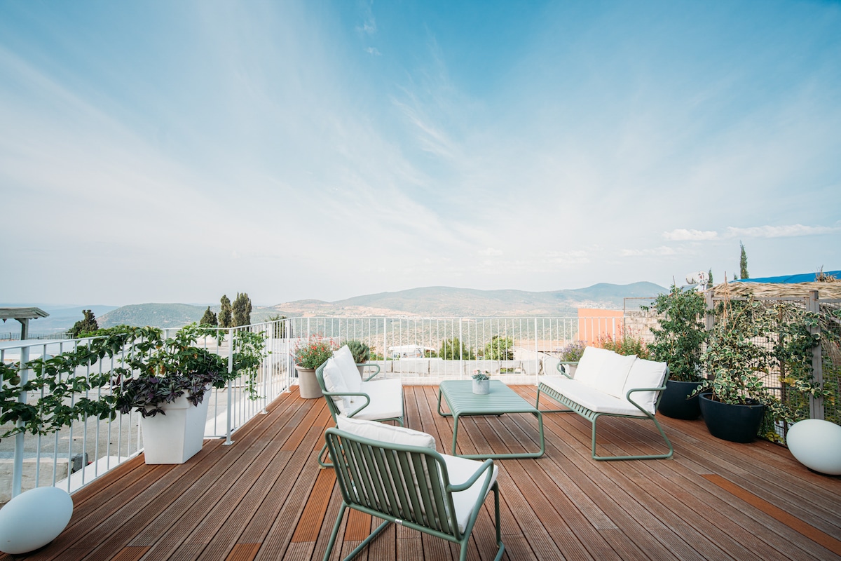 An outdoor deck features a cozy seating arrangement with modern chairs and a glass table. Potted plants add greenery, while a panoramic view of the surrounding hills and valleys is visible under a clear sky.