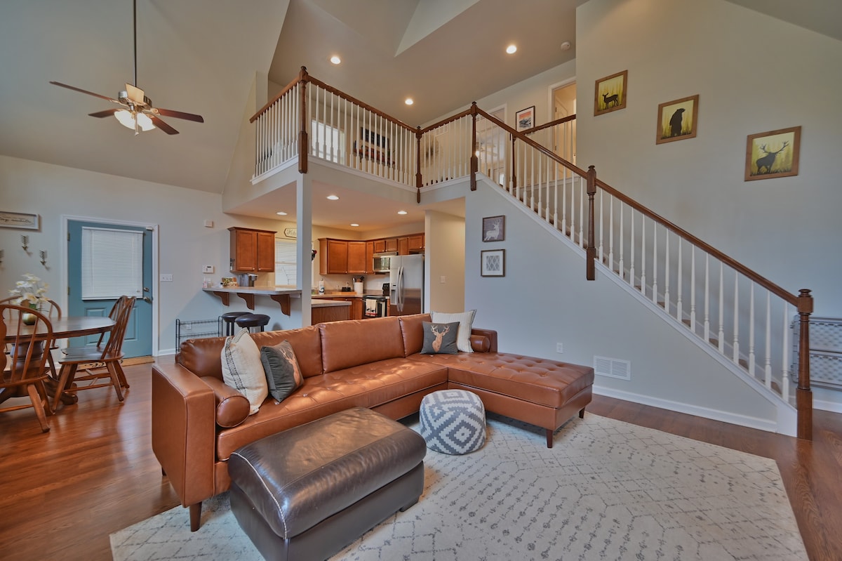A spacious living room is highlighted by a brown sectional sofa and a matching ottoman. High ceilings create an airy feel, with well-lit areas from large windows. The open layout reveals a dining area in the background and a staircase leading to an upper loft, enhancing the home’s inviting atmosphere.