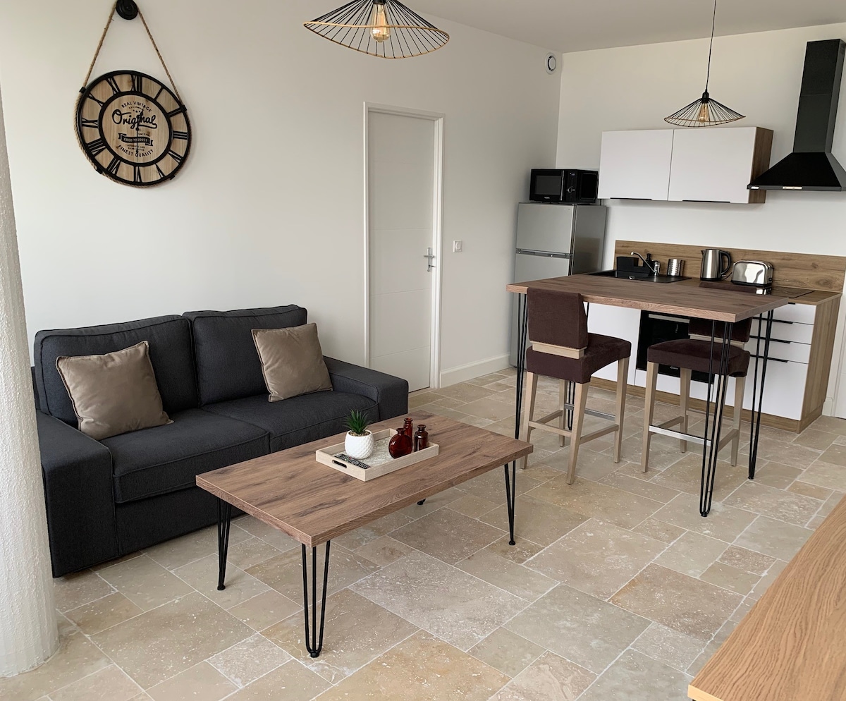 A bright living area is equipped with a comfortable dark sofa and a wooden coffee table. A dining space features two high-backed chairs and a tabletop. Modern kitchen appliances, including a refrigerator, are visible in the background, with warm light illuminating the space.