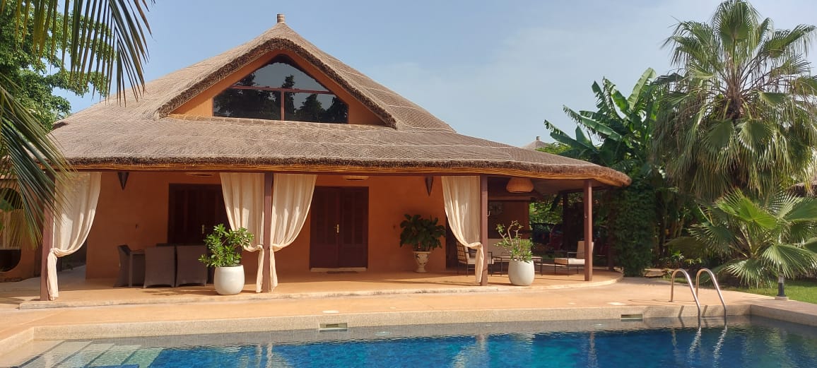 A serene villa with a thatched roof is depicted, featuring large windows that invite natural light. The patio area is equipped with comfortable seating and surrounded by lush greenery, leading to a private swimming pool with clear water, enhancing the tranquil outdoor atmosphere.