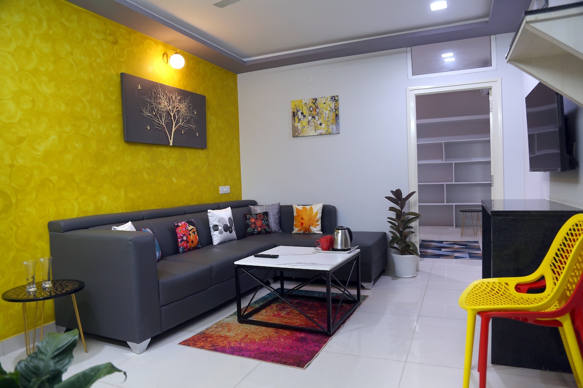 The living area features a modern gray sofa adorned with decorative cushions. A coffee table adds functionality, complemented by a colorful rug. The walls are painted in vibrant yellow, and there is a plant beside the sofa. Light is provided by an overhead fixture, and a doorway leads to additional spaces.