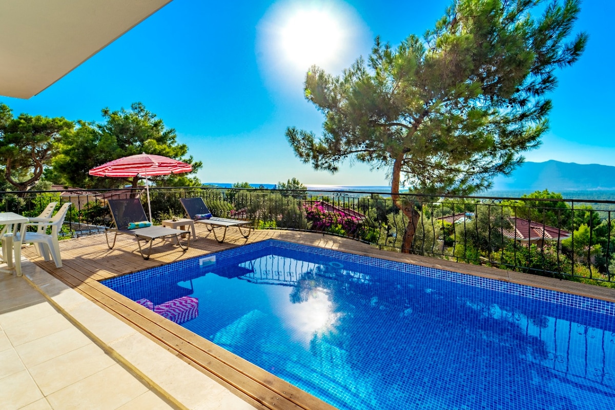 A private pool reflects the bright sun, surrounded by lush greenery. Sun loungers and a shaded umbrella are positioned on a wooden deck, providing a relaxing space to enjoy scenic views of the hills and Mediterranean coastline.