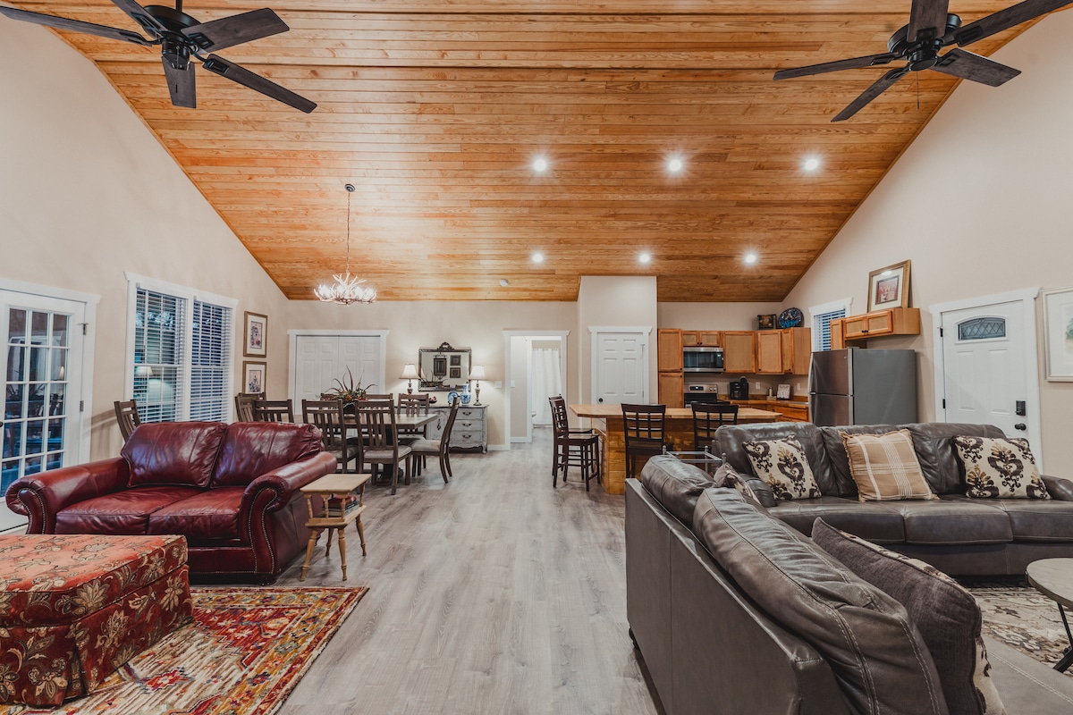 A spacious living area features high exposed wood ceilings and multiple ceiling fans. Comfortable seating is provided by leather sofas and a richly patterned ottoman. A dining area with a large table is visible, along with an open kitchen in the background, enhancing the communal feel of the space.