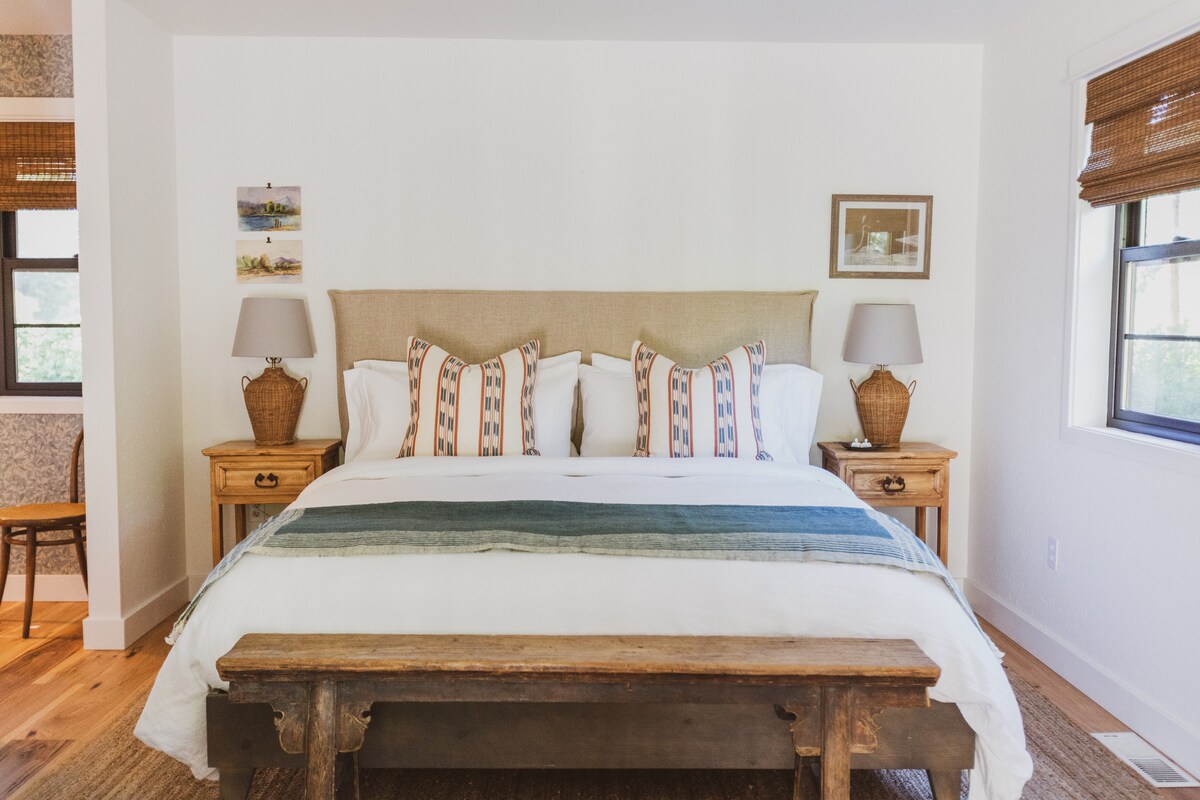 A cozy sleeping area features a spacious bed with decorative pillows arranged at the headboard. Two bedside tables with lamps flank the bed, and natural light enters through two windows adorned with woven shades.