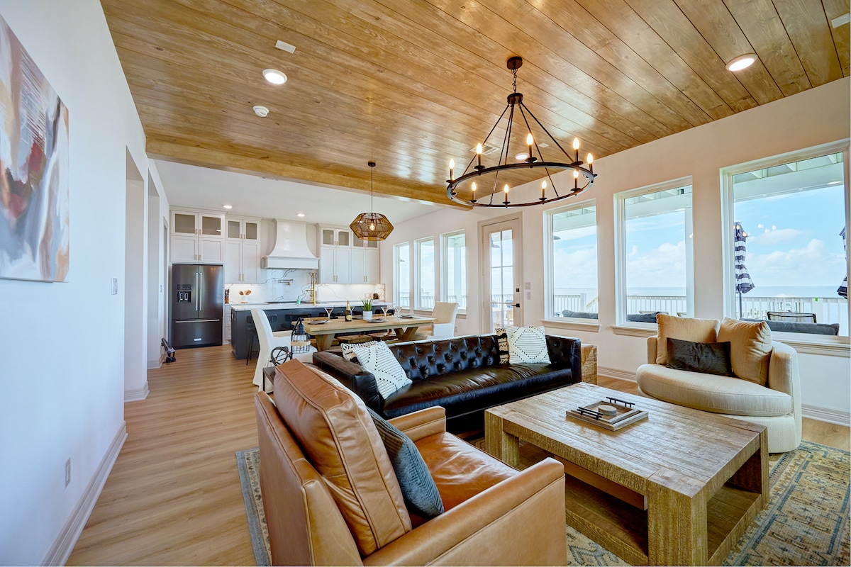 A spacious living area features a blend of modern and rustic design elements, highlighted by a wooden ceiling and large windows offering extensive views. Comfortable seating includes black and tan sofas arranged around a wooden coffee table, inviting relaxation and conversation.