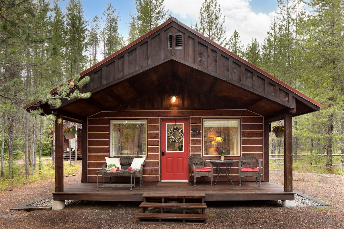 The cabin's rustic exterior features a large covered porch adorned with comfortable seating. Red front door is framed by two windows, while surrounding trees enhance the secluded feel. Wooden siding and a sloped roof complete the inviting design.