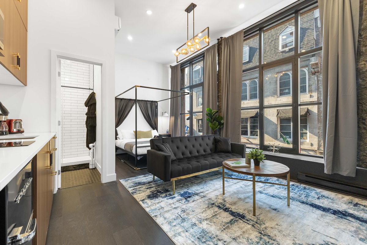 A spacious living area is presented, featuring large windows that allow natural light to filter in. A comfortable dark sofa and a round coffee table are centered on an area rug. A four-poster bed is visible in the background, separated by curtains.