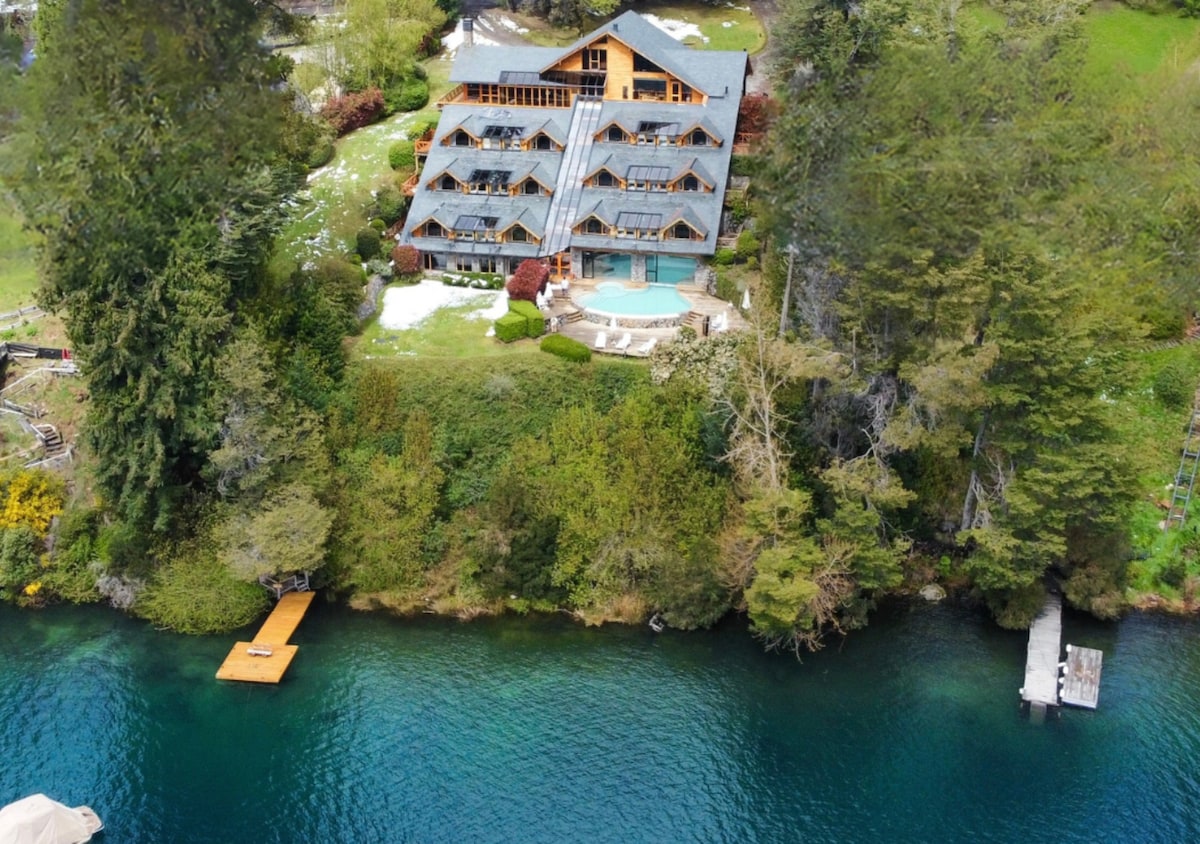 An aerial view reveals a multi-level lodge poised gracefully on a lush green hillside, overlooking clear blue waters. The building features a distinctive wooden design and is surrounded by trees. A wooden dock extends into the tranquil lake, enhancing the natural setting.