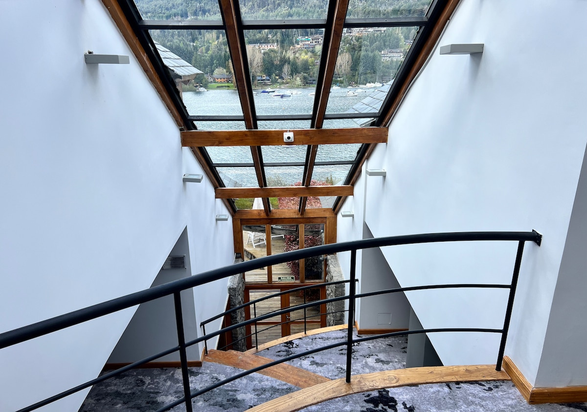 A staircase is visible, framed by large glass panels allowing natural light to fill the space. The warm wooden railing contrasts with the modern design, leading the eye towards a stunning view of the lake and surrounding landscape through expansive windows.