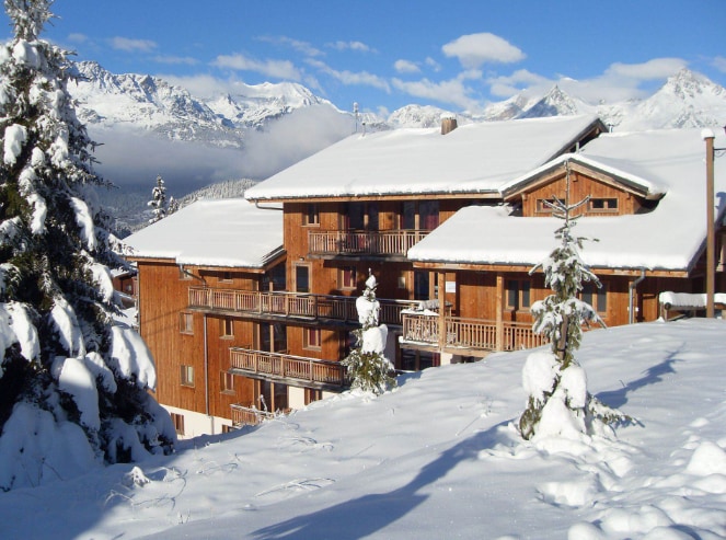 A three-story wooden chalet surrounded by a blanket of fresh snow. Balconies adorned with wooden railings overlook a winter landscape featuring distant mountains under a bright blue sky. Evergreen trees are dusted with snow, enhancing the serene alpine atmosphere.
