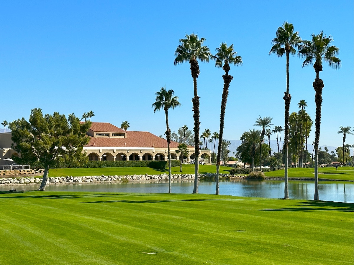 A well-maintained golf course is depicted with lush green grass, bordered by palm trees. A serene body of water reflects the clear blue sky, with a resort-style building visible in the background, offering a tranquil view from the grounds.