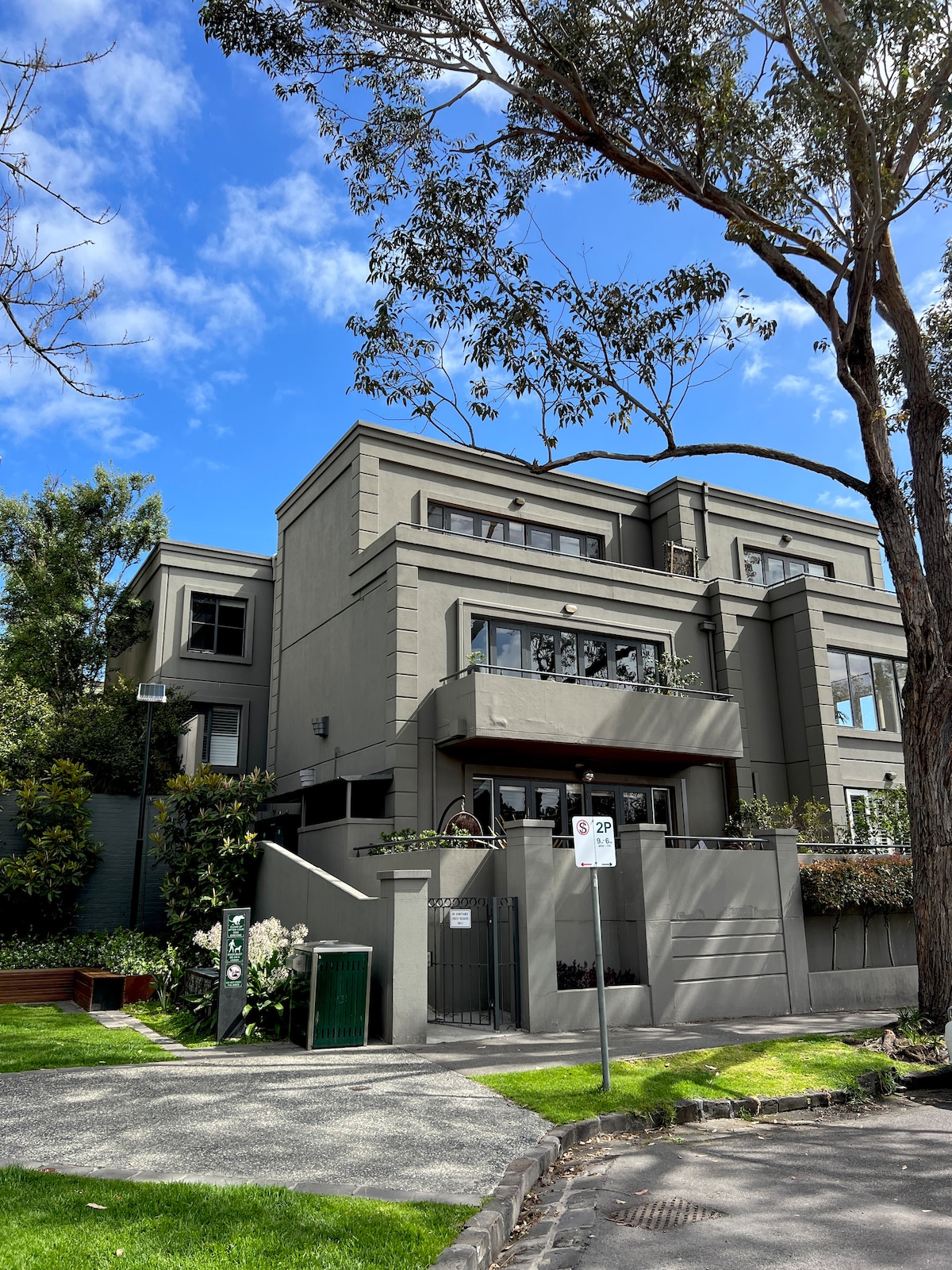 The exterior of a modern building is depicted, showcasing a blend of stone and large glass windows. Lush greenery surrounds the property, with trees and manicured lawns enhancing the inviting entrance. A bright blue sky with a few clouds serves as the backdrop.