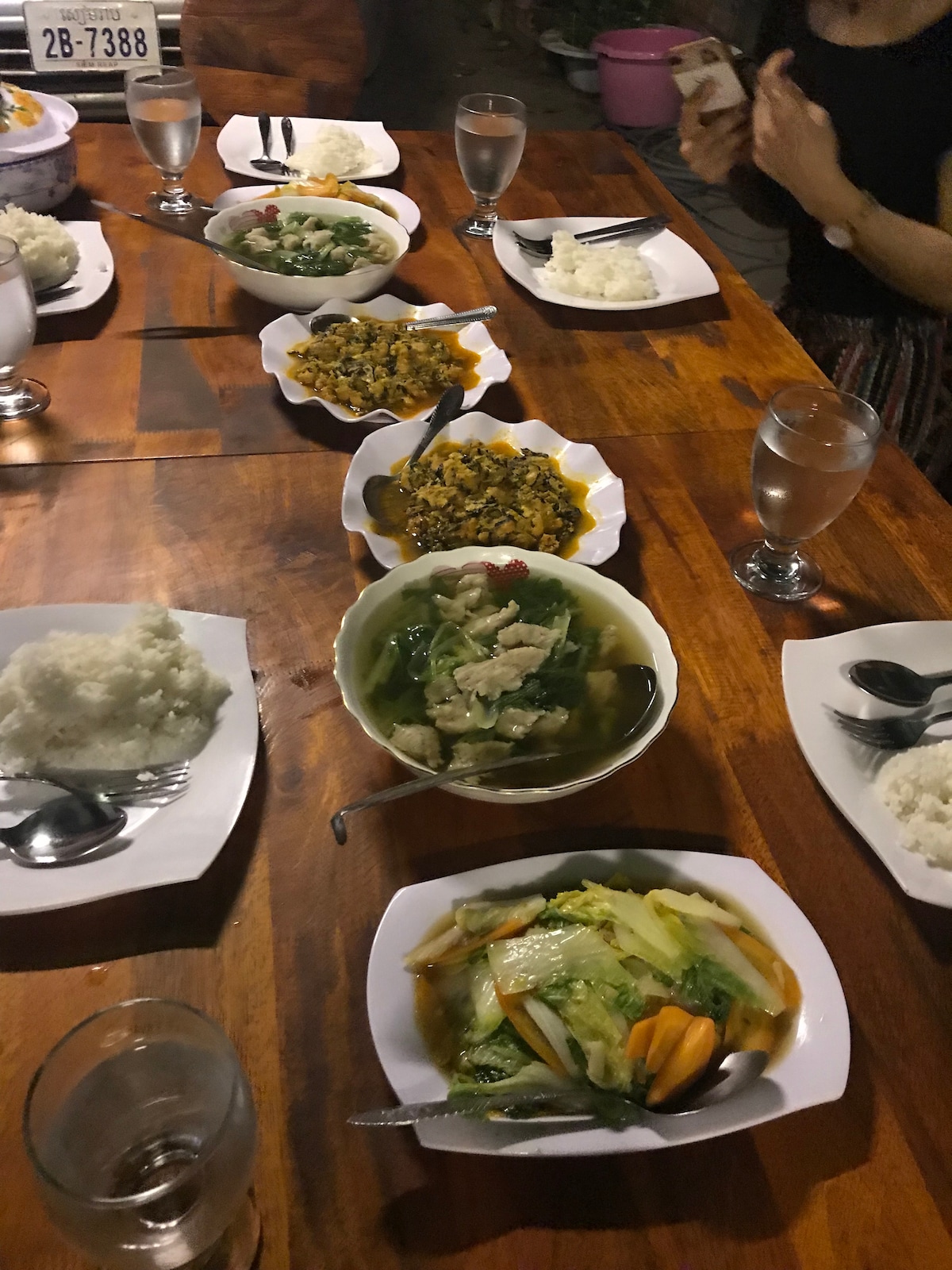 A wooden dining table is set with various dishes, including rice, vegetables, and savory meats served in white bowls. Several drinking glasses filled with water are visible, along with utensils arranged neatly. The atmosphere suggests a shared dining experience among guests.