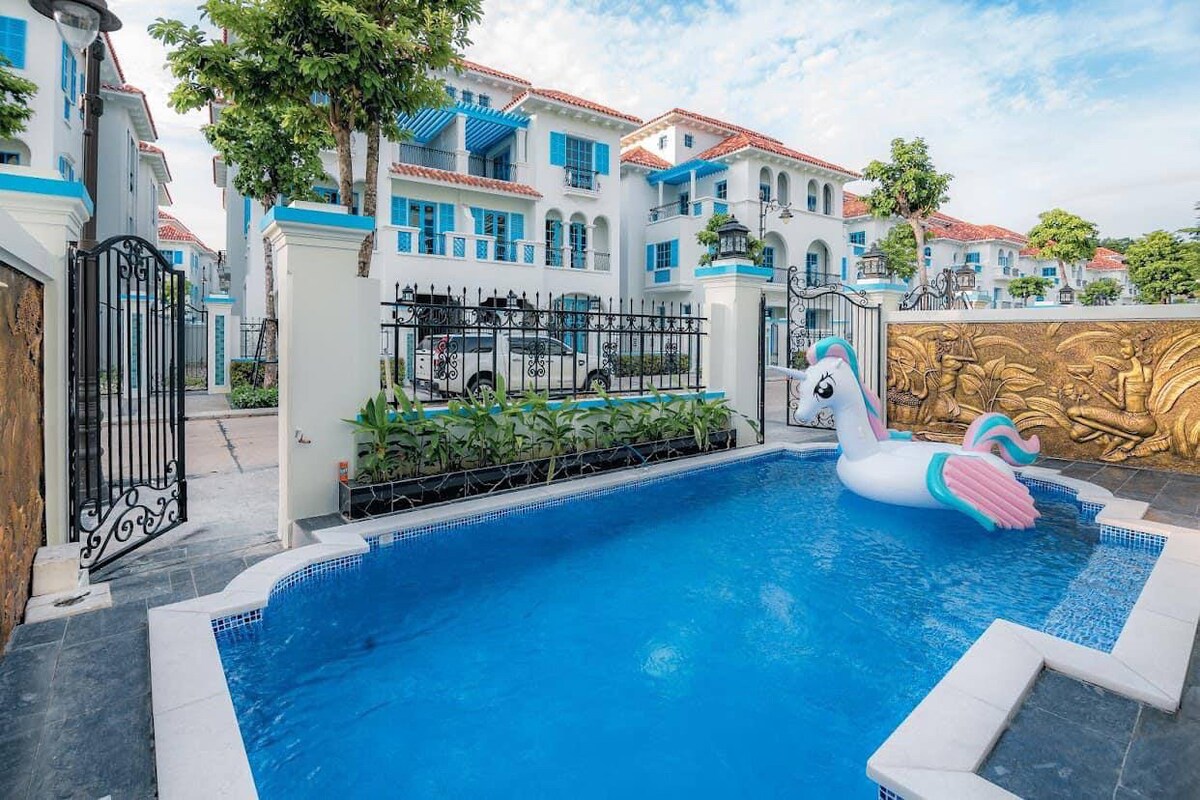 An inviting outdoor pool area is framed by manicured gardens and stylish architecture. A whimsical unicorn float rests in the clear water, creating a playful element. The villa’s exterior features a blend of white and blue elements, enhancing the serene atmosphere of the space.