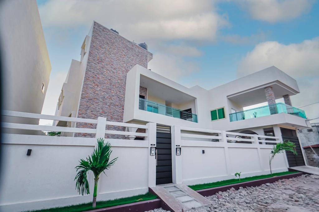 The exterior of the property showcases a modern design with a mix of stone and smooth surfaces. A neat pathway leads to the entrance, surrounded by well-maintained greenery, including a palm plant. Large balcony spaces are visible on the upper levels.
