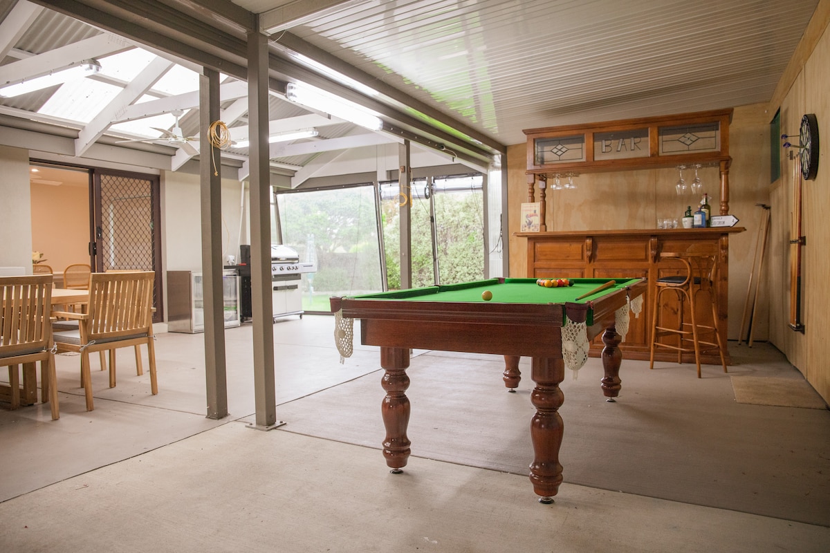 An indoor games area features a polished billiard table centered on a concrete floor. Natural light enters through large glass doors, illuminating a wooden bar with stools and a nearby outdoor dining set. A BBQ grill is visible in the background.