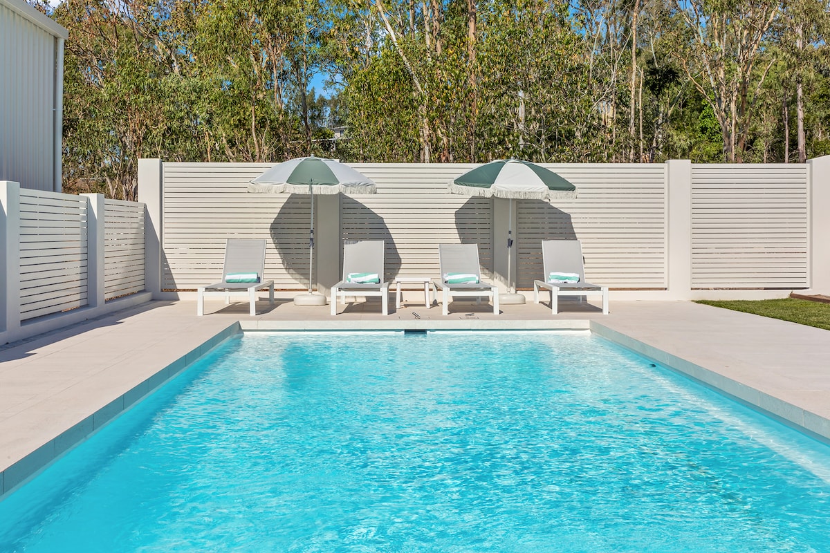 A private pool area is surrounded by a light-colored deck and features three lounge chairs beneath matching umbrellas. The pool's clear blue water reflects sunlight, and a green lawn is visible in the background, providing a serene outdoor setting.