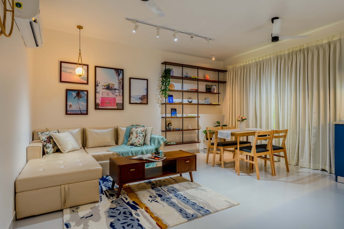 A bright living area features a spacious sofa, a wooden coffee table, and a dining set for four. Shelving units showcase decorative items and plants. Large windows with sheer curtains let in natural light, enhancing the modern decor with framed art on the walls.