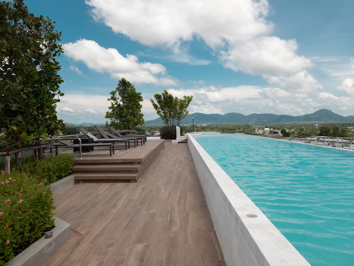 A panoramic pool area is featured on the rooftop, showcasing an elongated infinity pool that blends with the skyline. Lounge chairs are positioned along the wooden deck, surrounded by greenery and offering views of distant mountains under a cloudy sky.