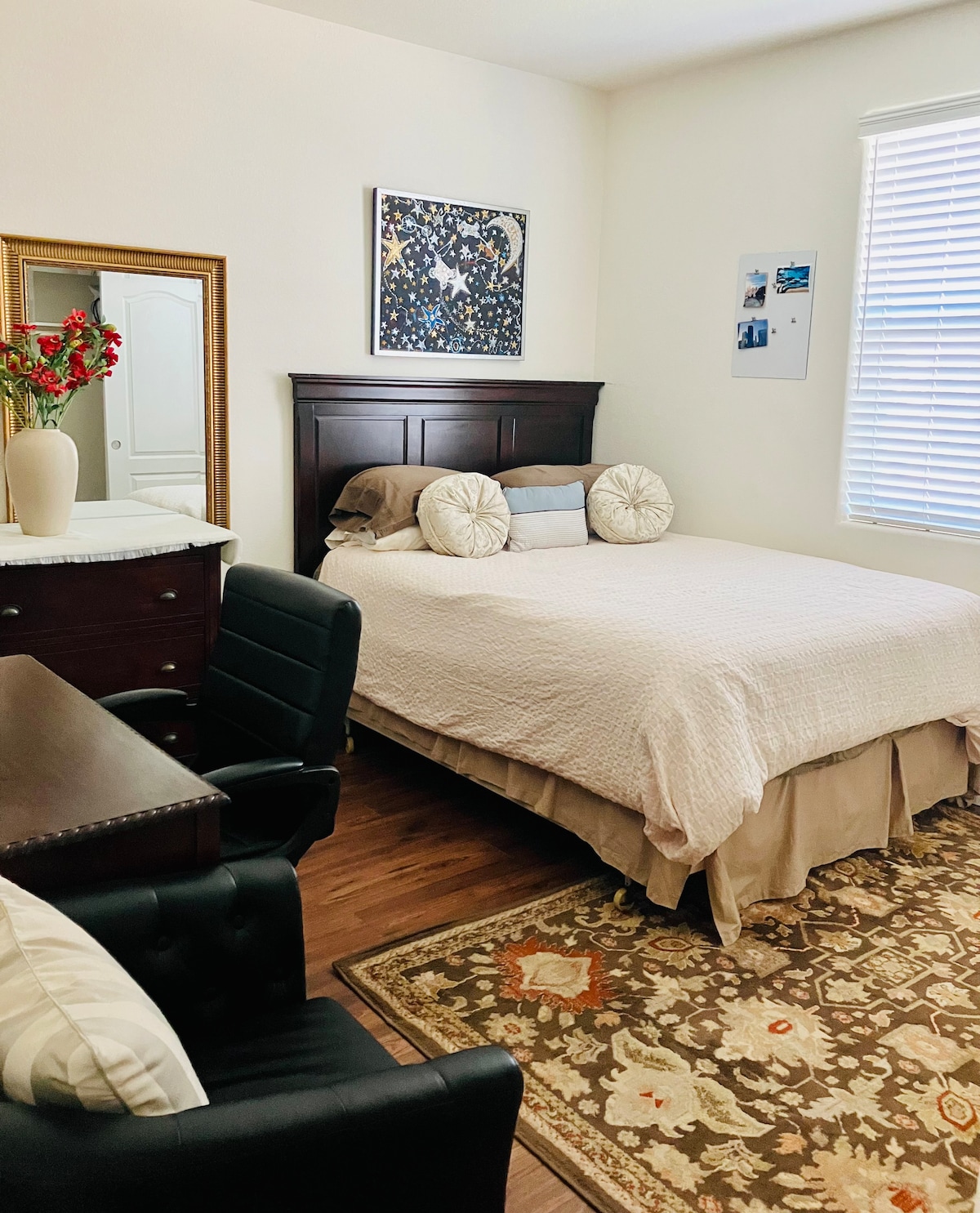 A well-appointed bedroom is featured, showcasing a spacious bed with soft linens and two decorative pillows. A wooden desk and two chairs provide a workspace, while a mirror reflects natural light. The room is complemented by a patterned area rug and wall art.