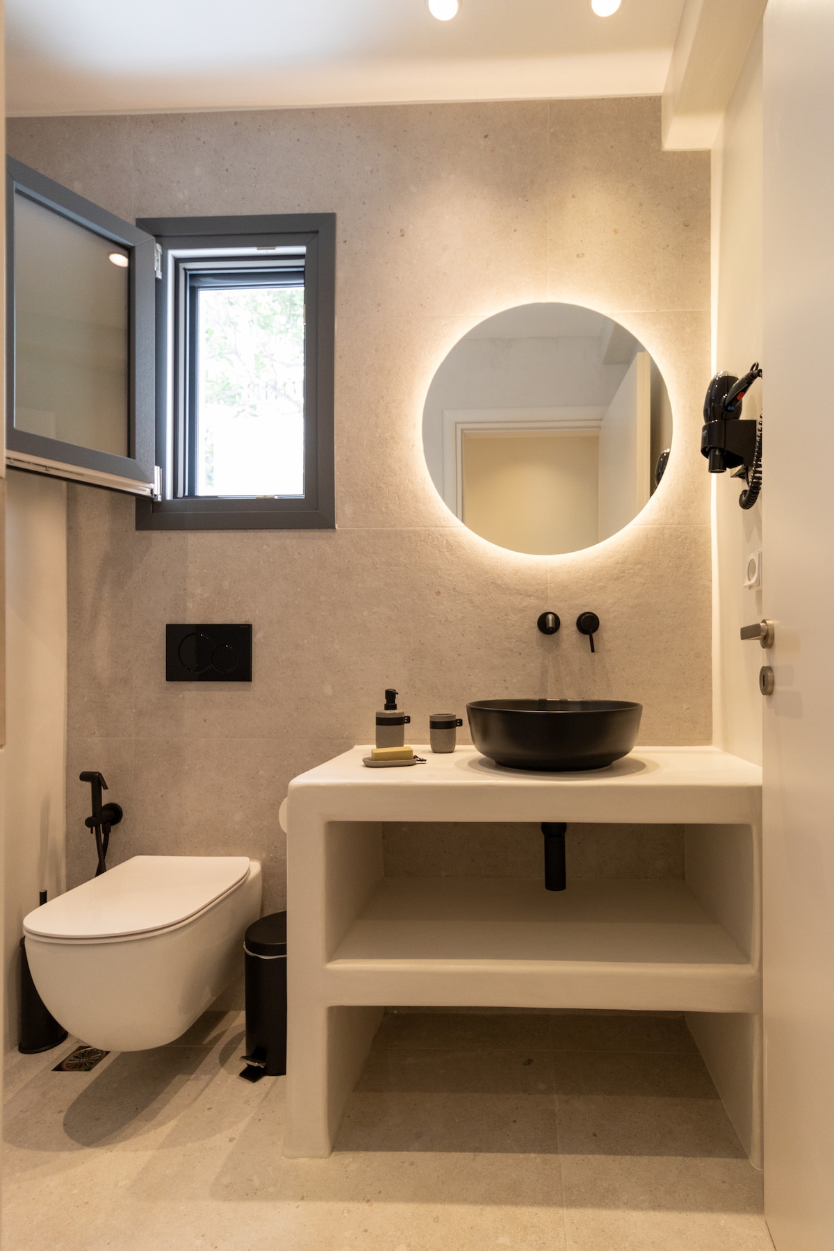 The bathroom features a minimalist design with light-colored stone walls. A round mirror is mounted above a sleek black basin atop a simple countertop. Natural light enters through a small window, and a modern toilet is located beside the sink area.