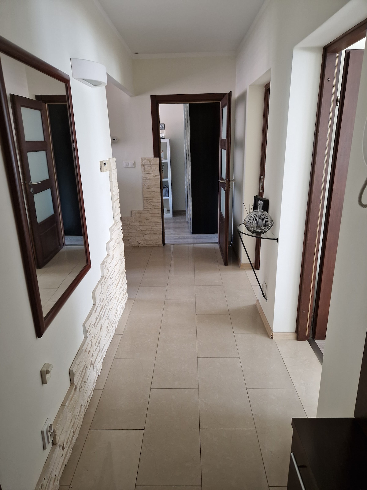 A hallway is shown with tiled flooring and light-colored walls. Two mirrors are positioned on the left side, reflecting the space. Several wooden doors lead to adjoining rooms, adding depth to the area. Small decorative stone accents are visible on the wall.